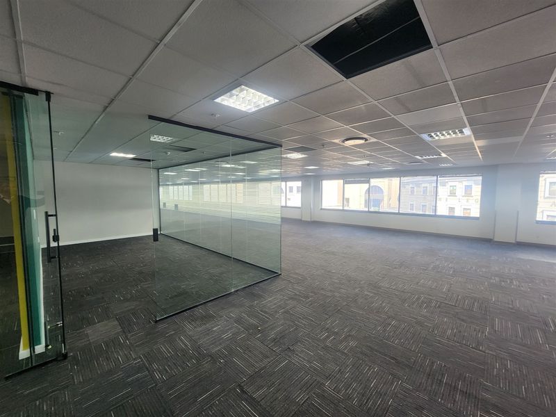 Spacious open-plan office area featuring carpet tiles, a glass-partitioned meeting room, and large windows providing ample natural light.