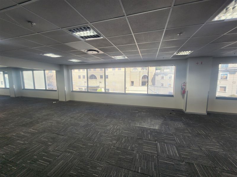Spacious, empty open-plan office area featuring carpet tiles, suspended ceilings with recessed lighting, and large windows providing ample natural light.