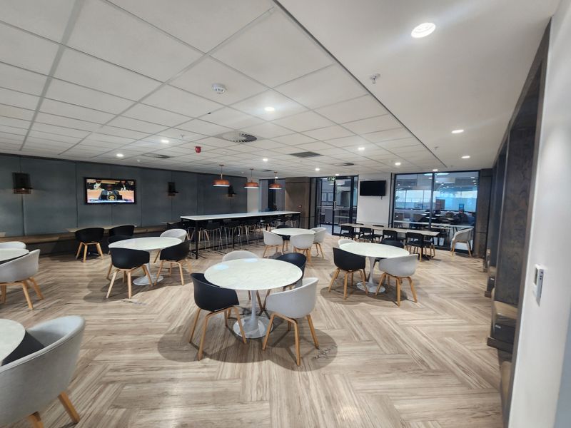 Modern open-plan office breakout area featuring herringbone flooring, suspended ceiling, recessed lighting, and stylish upholstered wall paneling with integrated bench seating.