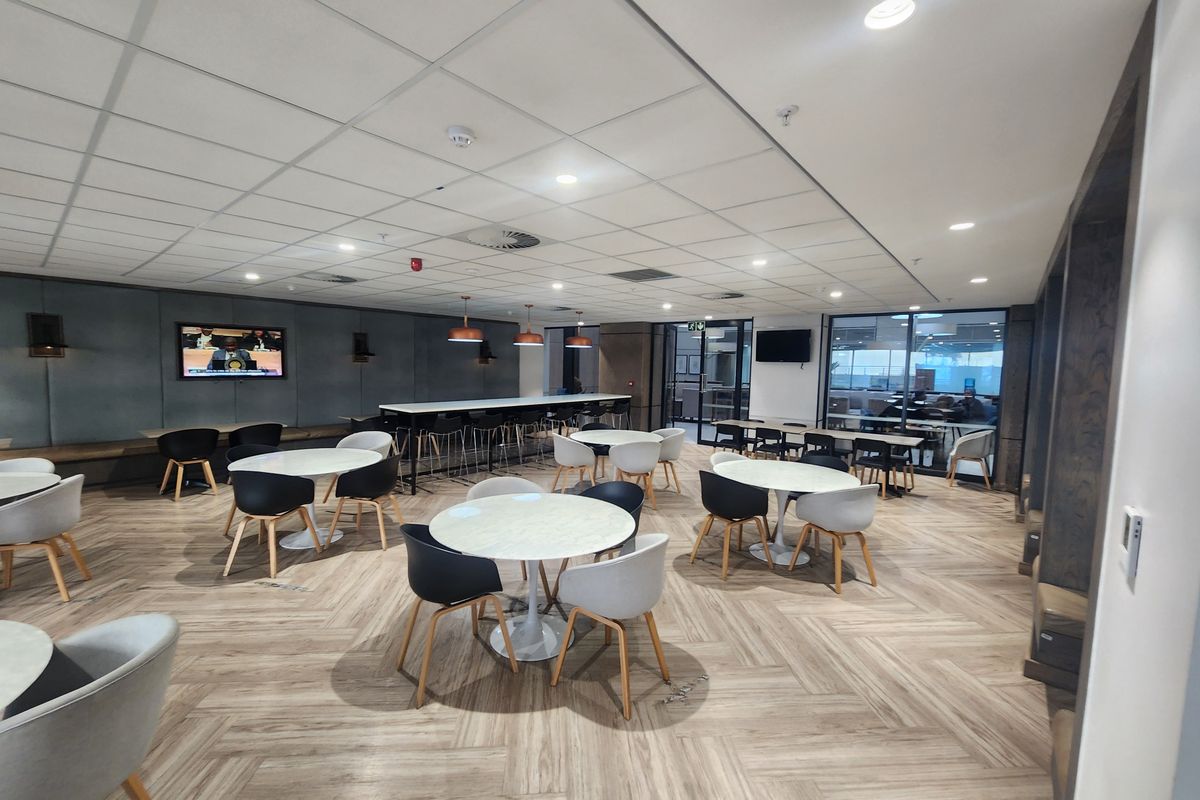 Modern open-plan office breakout area featuring herringbone flooring, suspended ceiling, recessed lighting, and stylish upholstered wall paneling with integrated bench seating.
