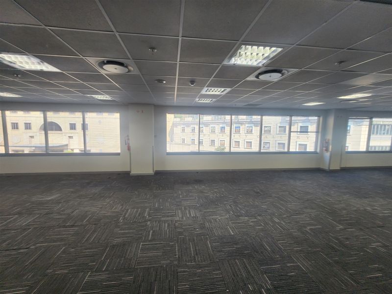 An empty, open-plan office space featuring suspended ceilings, recessed lighting, and large windows providing natural light and urban views.