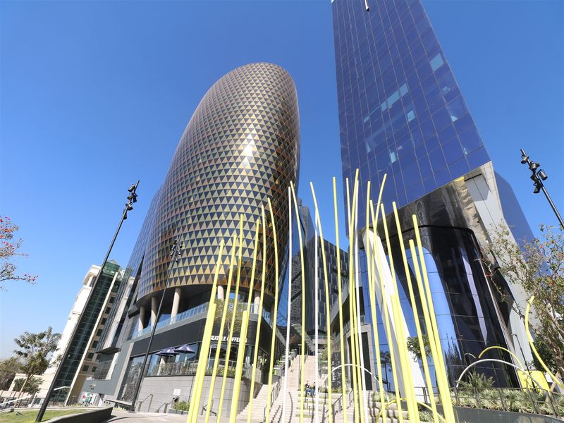 A low-angle view of a modern office complex featuring a distinctive gold-patterned curved tower and adjacent glass skyscraper with a landscaped pedestrian plaza.