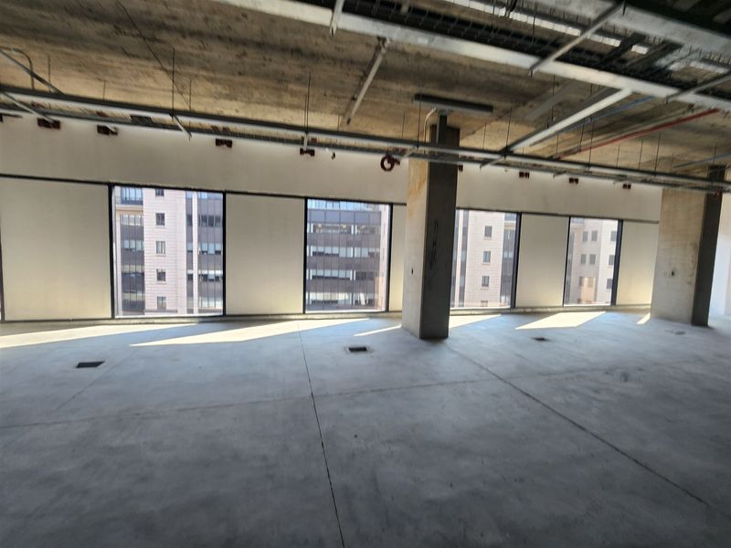 An open-plan office space in shell condition, featuring exposed concrete ceilings, large windows with city views, and raw concrete flooring.