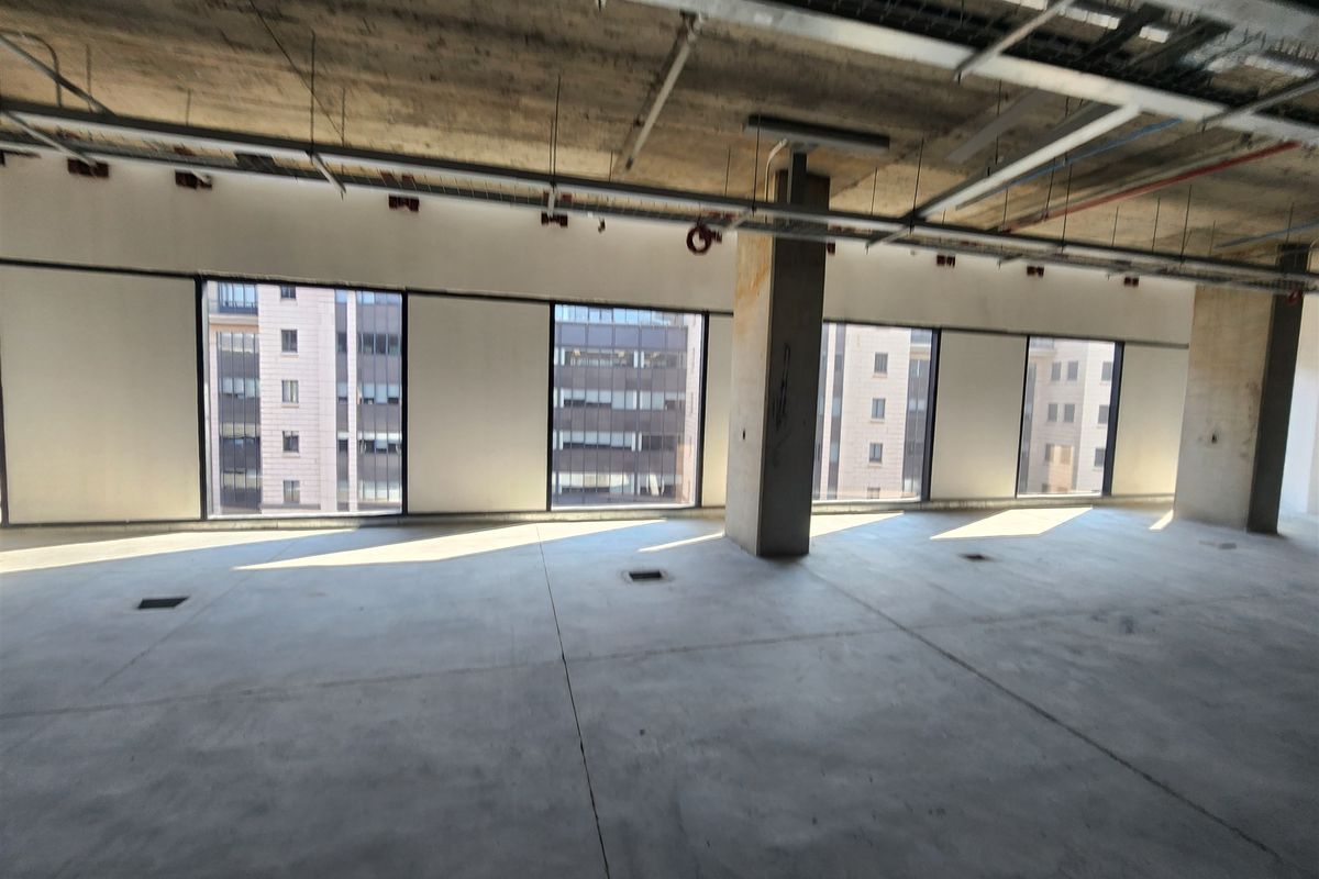 An open-plan office space in shell condition, featuring exposed concrete ceilings, large windows with city views, and raw concrete flooring.