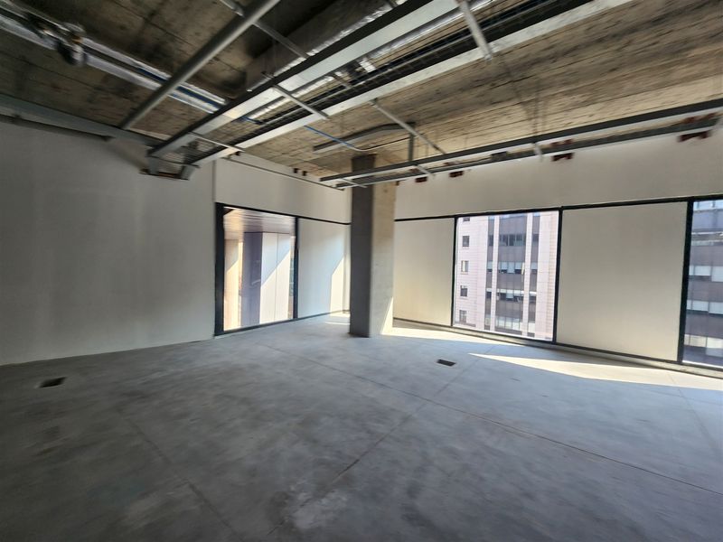 An open-plan office space in shell condition, featuring exposed concrete ceilings, large windows with city views, and raw concrete flooring.