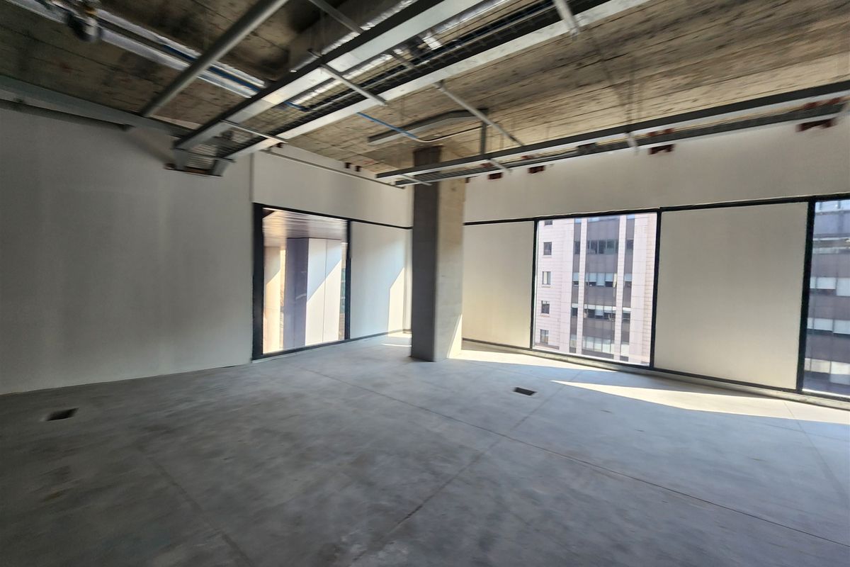 An open-plan office space in shell condition, featuring exposed concrete ceilings, large windows with city views, and raw concrete flooring.
