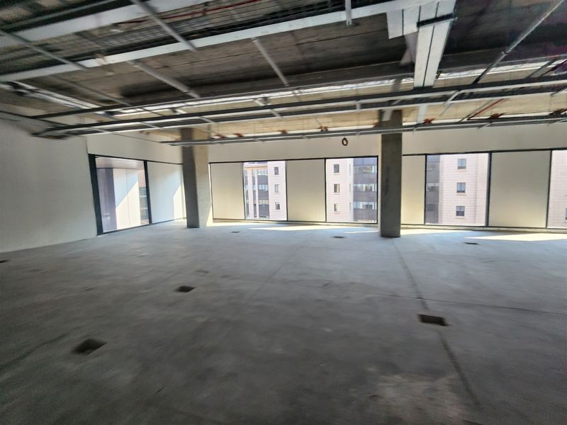 An open-plan office space in shell condition featuring exposed ceiling infrastructure, concrete flooring, and large windows providing natural light.