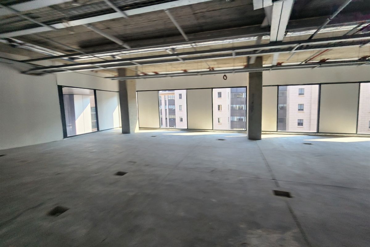An open-plan office space in shell condition featuring exposed ceiling infrastructure, concrete flooring, and large windows providing natural light.
