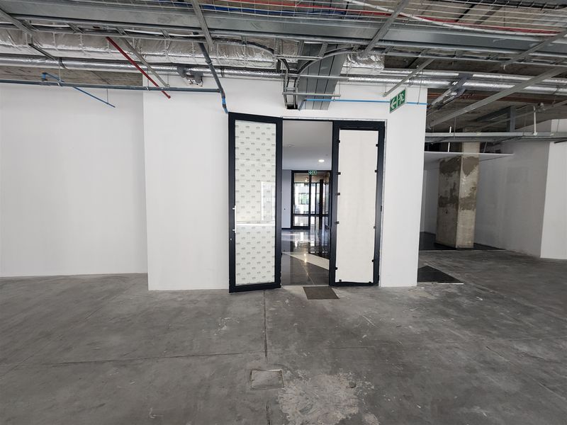 An unfinished office space featuring exposed ceiling services, concrete flooring, and double glass doors, currently in a shell condition ready for fit-out.