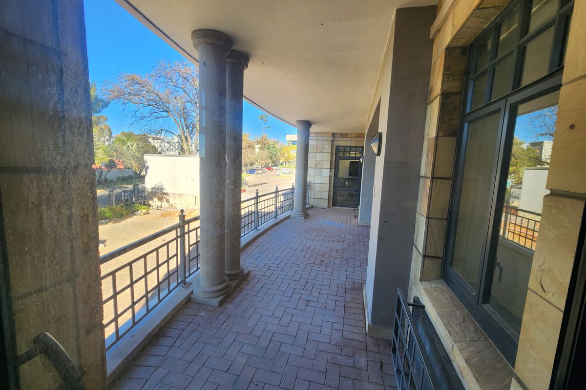 A covered outdoor balcony featuring stone columns, brick paving, and metal railings, providing an open-air transition space for the office building.