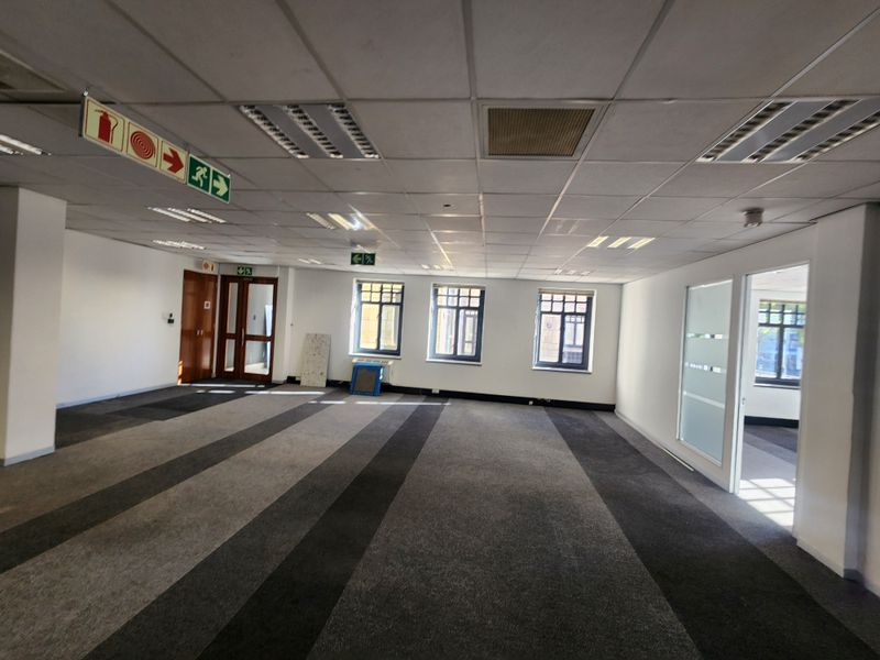 Spacious, open-plan office area featuring a suspended ceiling, recessed lighting, carpeted flooring, and glass partition doors, providing a versatile workspace.