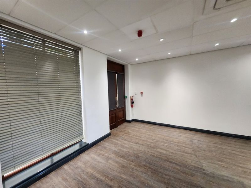 A clean, vacant office space featuring laminate flooring, a suspended ceiling with recessed lighting, and large windows fitted with venetian blinds.