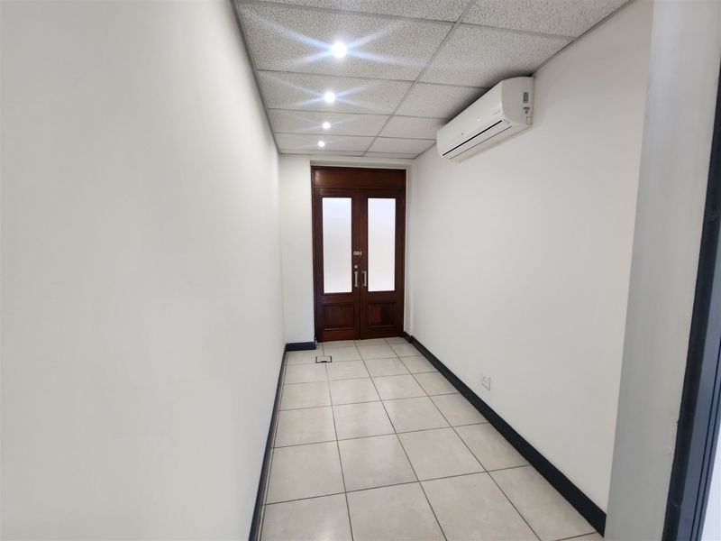 A clean, empty office space featuring tiled flooring, a suspended ceiling with recessed lighting, and a wall-mounted air conditioning unit.