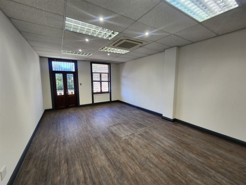 A vacant office space featuring laminate flooring, a suspended ceiling with integrated lighting, a cassette air conditioning unit, and timber-framed doors with security gates.