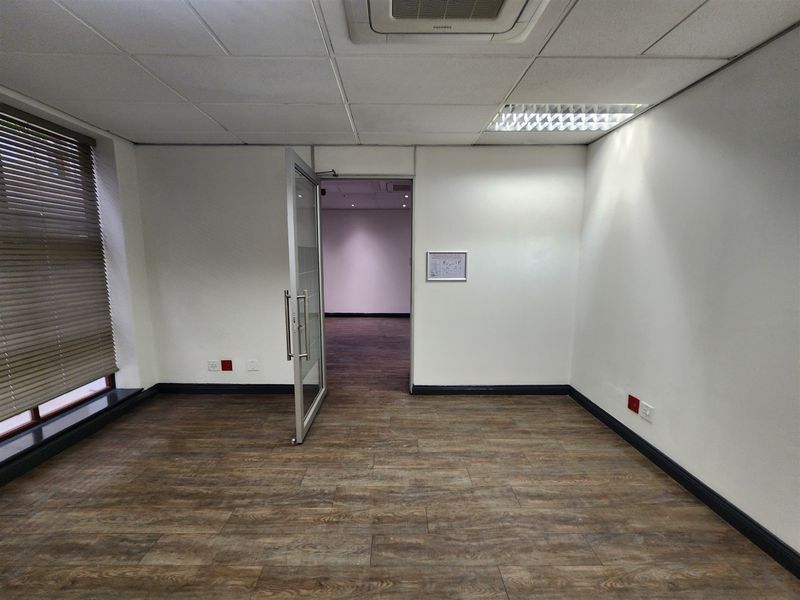 A clean, vacant office space featuring wood-look flooring, a suspended ceiling with recessed lighting, a cassette air conditioning unit, and a glass entrance door.