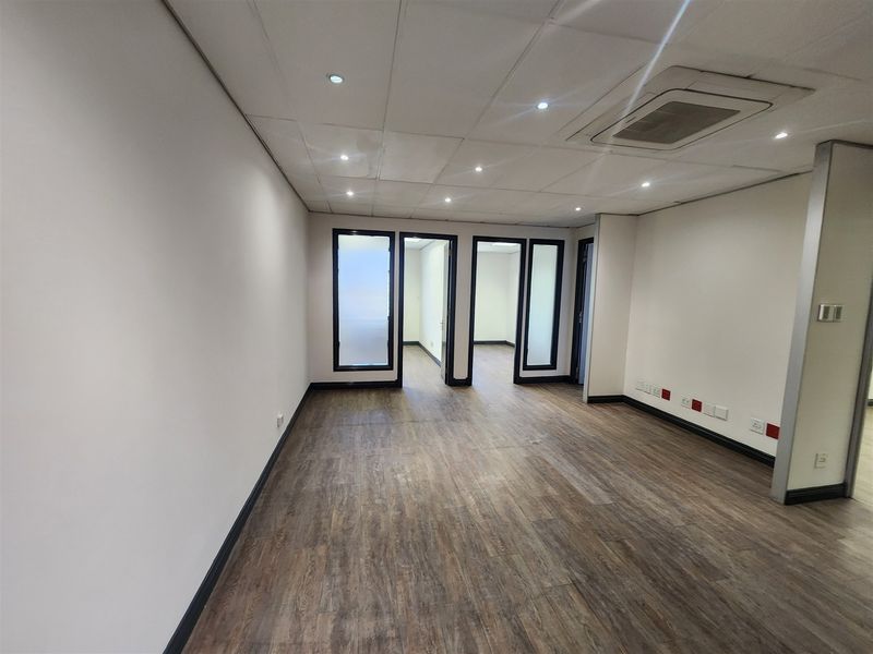 A bright, vacant office space featuring modern laminate flooring, a suspended ceiling with recessed lighting, and glass-paneled interior doors.