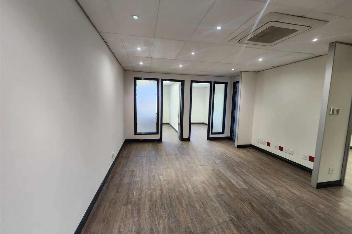 A bright, vacant office space featuring modern laminate flooring, a suspended ceiling with recessed lighting, and glass-paneled interior doors.