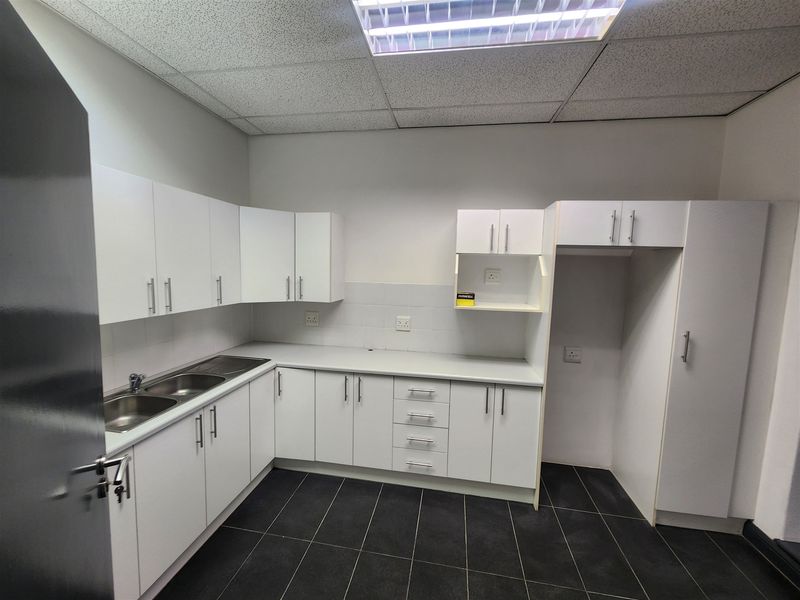 A clean, functional office kitchenette featuring white built-in cabinetry, a stainless steel double sink, dark tiled flooring, and recessed ceiling lighting.