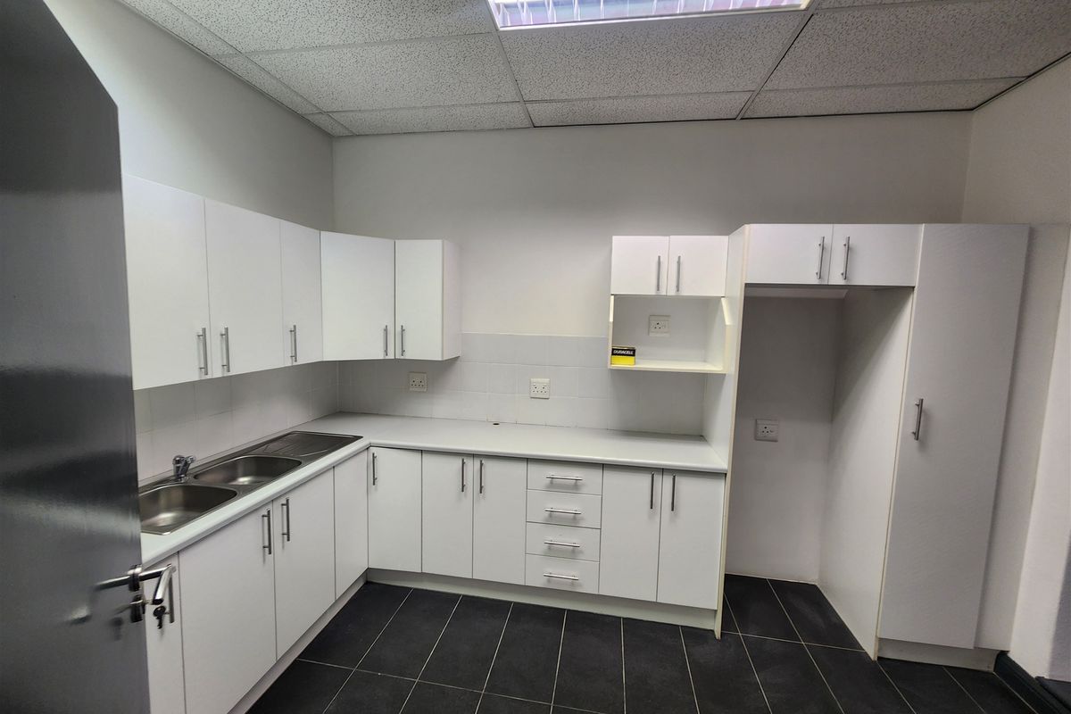 A clean, functional office kitchenette featuring white built-in cabinetry, a stainless steel double sink, dark tiled flooring, and recessed ceiling lighting.