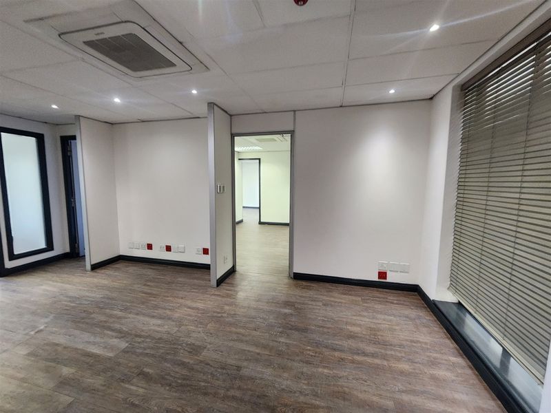 A clean, empty office space featuring laminate flooring, a suspended ceiling with recessed lighting, integrated air conditioning, and large windows with venetian blinds.