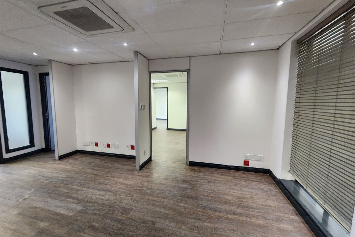 A clean, empty office space featuring laminate flooring, a suspended ceiling with recessed lighting, integrated air conditioning, and large windows with venetian blinds.