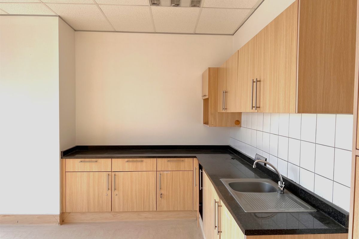 A clean, functional office kitchenette featuring light wood-grain cabinetry, dark stone countertops, a stainless steel sink, and a tiled splashback.