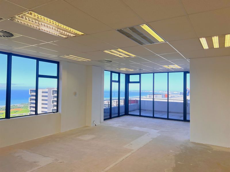 An empty, open-plan office space featuring large windows with panoramic coastal views, a suspended ceiling, and direct access to a balcony.