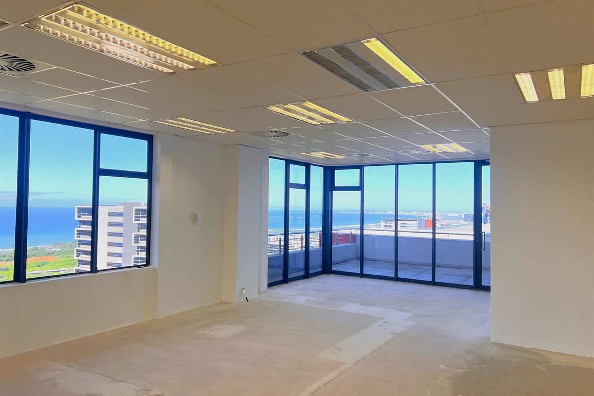 An empty, open-plan office space featuring large windows with panoramic coastal views, a suspended ceiling, and direct access to a balcony.
