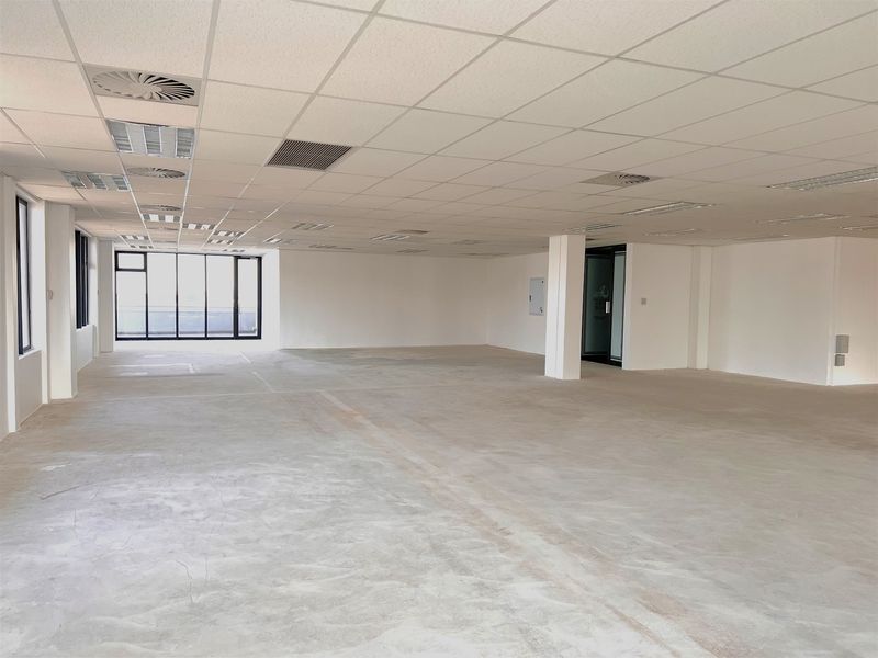 Spacious open-plan office area featuring a suspended ceiling, recessed lighting, air conditioning vents, and large windows with concrete flooring.