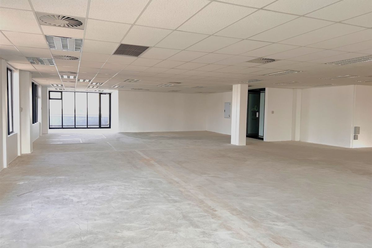 Spacious open-plan office area featuring a suspended ceiling, recessed lighting, air conditioning vents, and large windows with concrete flooring.
