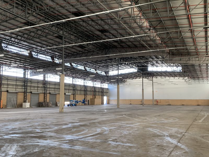 Expansive industrial warehouse space featuring high ceilings with exposed steel trusses, concrete flooring, multiple roller shutter doors, and integrated fire sprinkler systems.