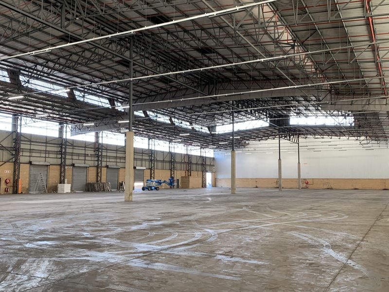 Expansive industrial warehouse space featuring high ceilings with exposed steel trusses, concrete flooring, and multiple roller shutter doors for efficient logistics access.