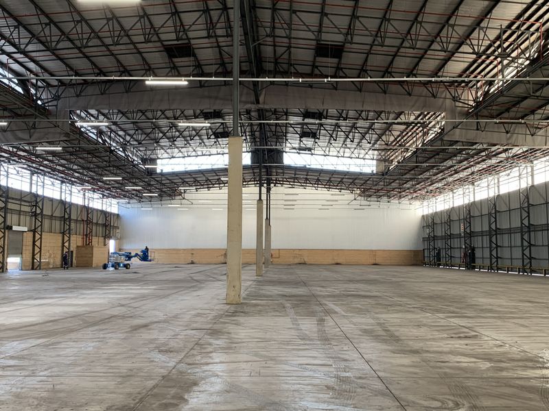 Expansive industrial warehouse space featuring high ceilings with exposed steel trusses, concrete flooring, and large roller shutter access points.