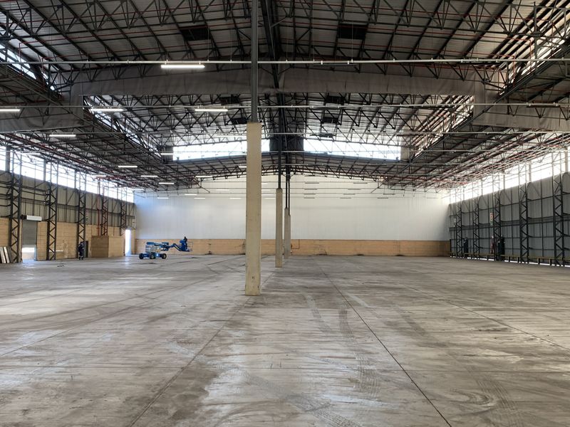 Large open-plan industrial warehouse space featuring high ceilings with exposed steel trusses, concrete flooring, and multiple roller shutter access points.