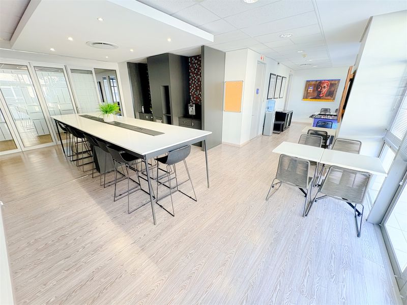 Modern open-plan office breakout area featuring a central high-top table, kitchenette station, and glass-partitioned meeting rooms with light wood-look flooring.