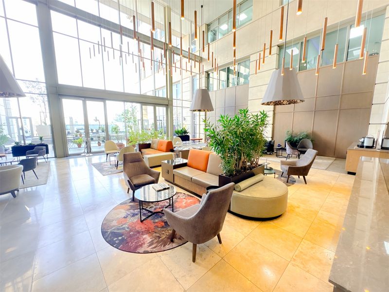 A spacious, modern office lobby featuring high ceilings, floor-to-ceiling windows, polished stone flooring, and contemporary pendant lighting.