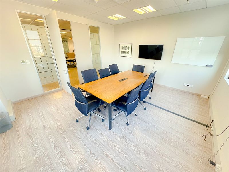 A professional office meeting room featuring a glass partition, wood-look laminate flooring, recessed lighting, and a clean, modern aesthetic.