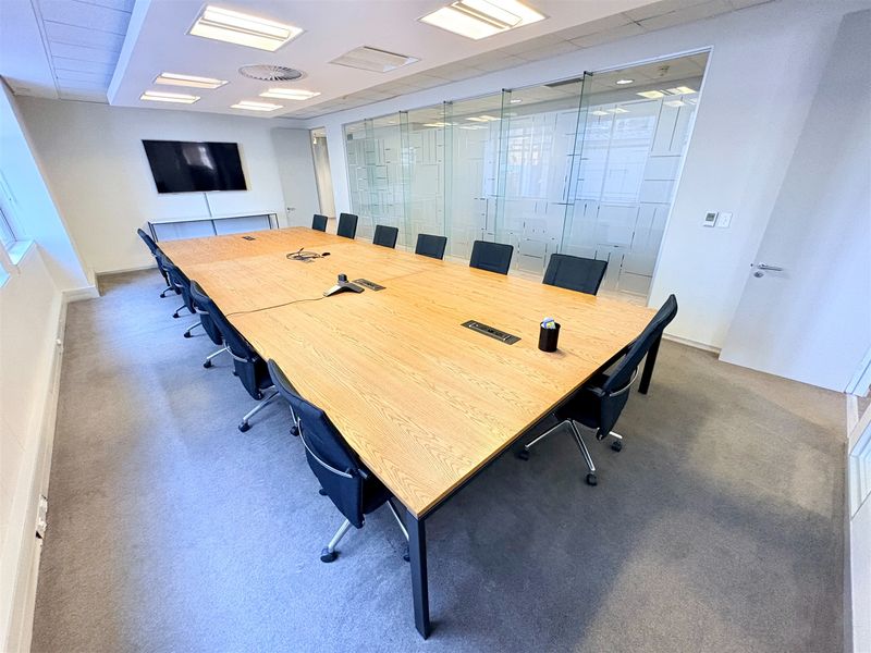 A professional boardroom featuring a large wooden conference table, glass partition walls, and recessed ceiling lighting, suitable for corporate meetings.