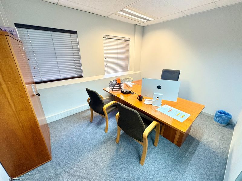 A functional office space featuring carpeted flooring, a suspended ceiling with recessed lighting, and windows fitted with venetian blinds.