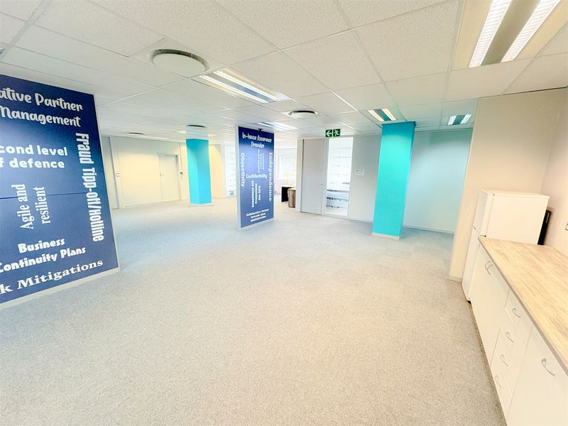 Spacious open-plan office area featuring carpeted flooring, suspended ceilings with recessed lighting, structural columns, and a compact built-in kitchenette unit.