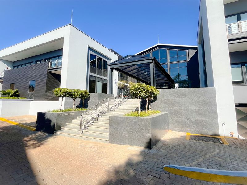 Modern office building exterior featuring a prominent glass entrance canopy, stone-clad retaining walls, and a paved approach with professional landscaping.