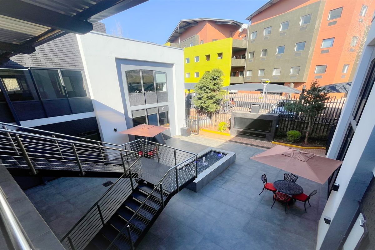 A modern, paved office courtyard featuring a central water feature, built-in braai area, and an external metal staircase with stainless steel railings.