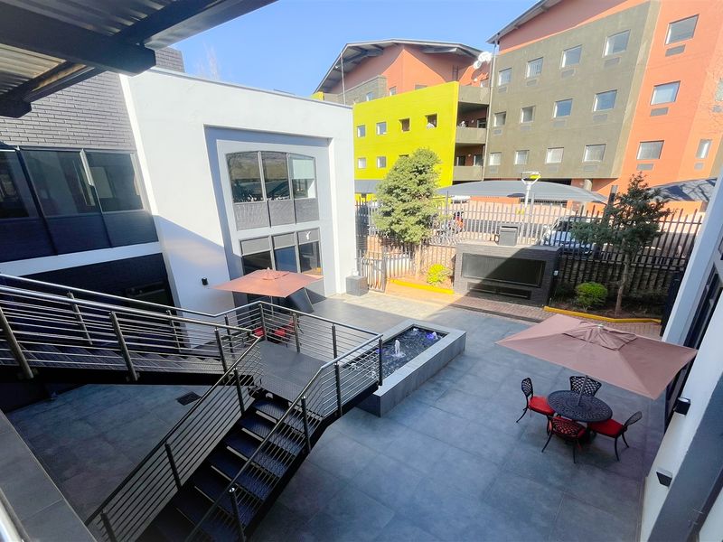 A modern, paved office courtyard featuring a central water feature, built-in braai area, and an external metal staircase with stainless steel railings.