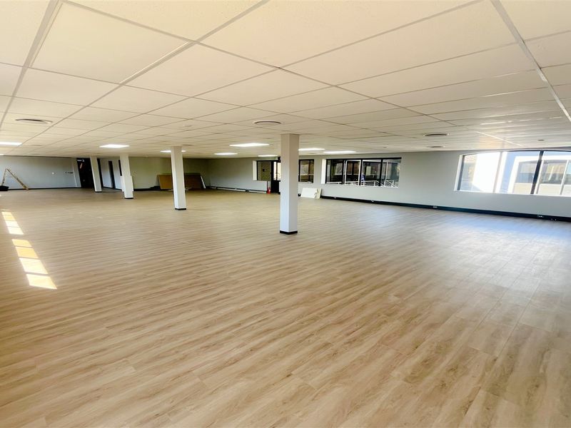 A spacious, open-plan office area featuring modern laminate flooring, a suspended ceiling with recessed lighting, and large windows providing ample natural light.