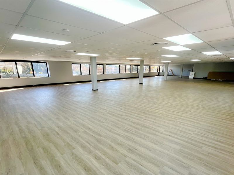 A spacious, open-plan office area featuring modern laminate flooring, suspended ceilings with recessed lighting, and large windows providing ample natural light.