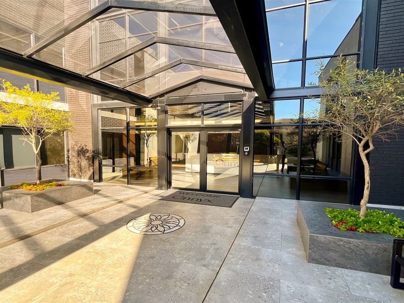 Modern office building entrance featuring a glass canopy, stone paved courtyard, and contemporary architectural glass facade.