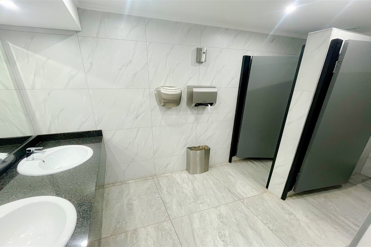 Modern office bathroom featuring a dual vanity with granite countertop, marble-look wall tiles, and private toilet cubicles.