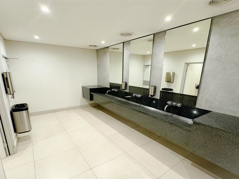 Modern commercial bathroom featuring a long granite vanity with integrated basins, wall-mounted faucets, large mirrors, and clean tiled flooring.