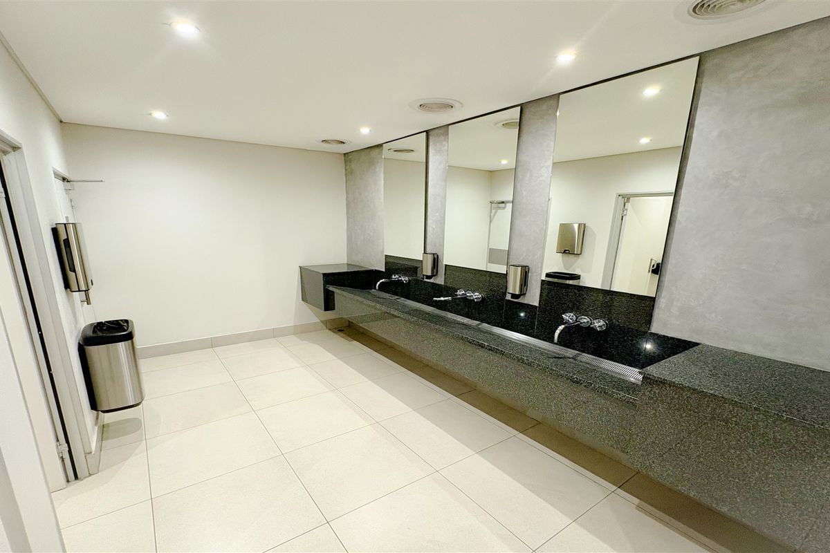 Modern commercial bathroom featuring a long granite vanity with integrated basins, wall-mounted faucets, large mirrors, and clean tiled flooring.
