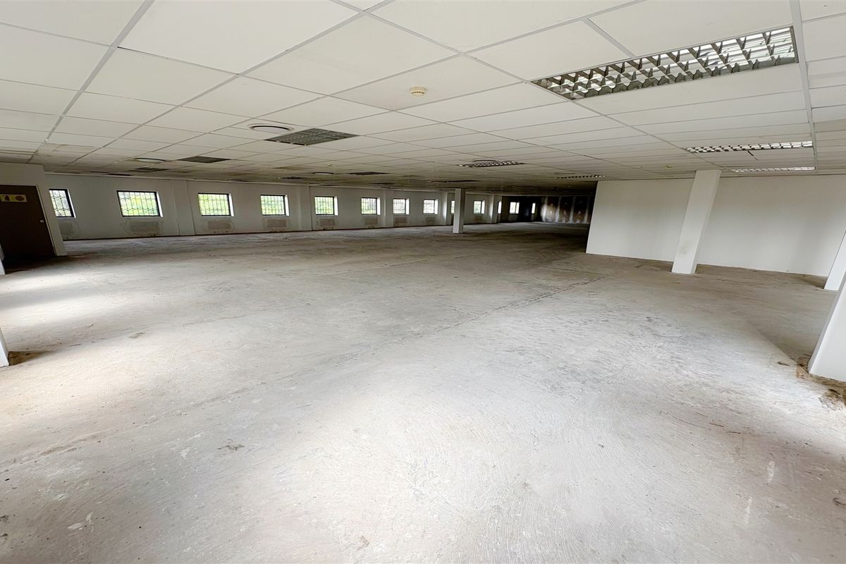 A large, open-plan office space featuring a suspended ceiling, recessed lighting, and security-barred windows, currently in a vacant, shell-like condition.